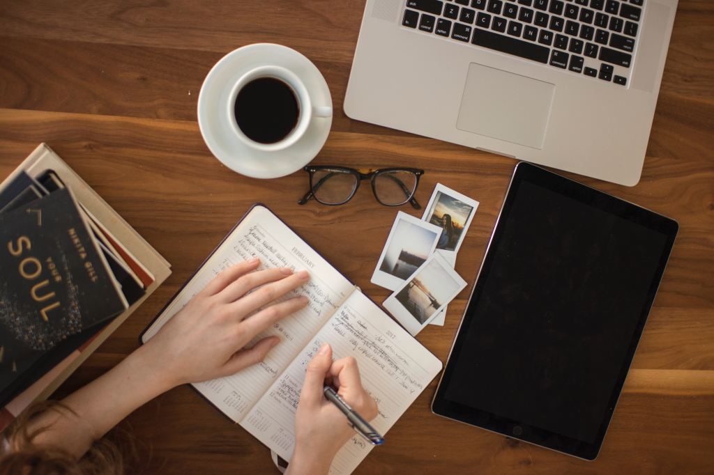 top view of a person writing on a notebook with a laptop, eye glasses, coffee, books and tablet